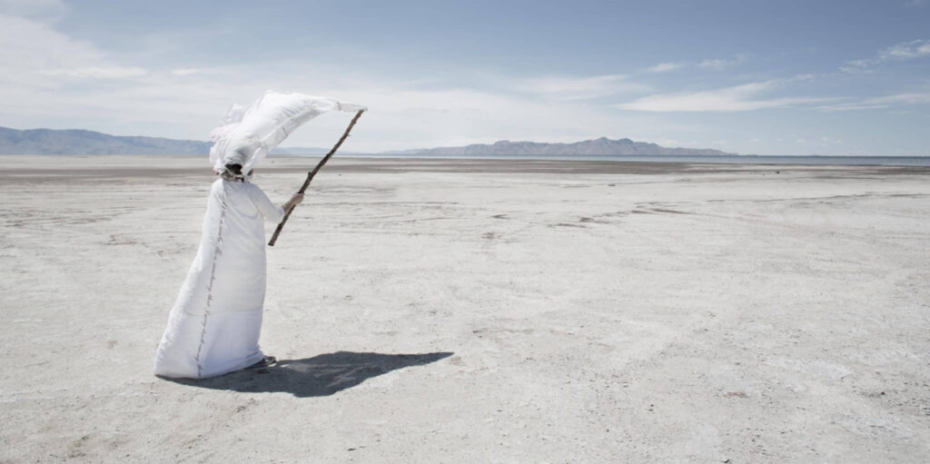 Prof. Beth Krensky walking along the salt flats dressed in a rob and headpiece attached to a stick.